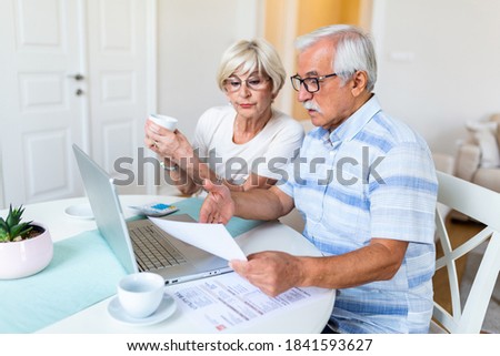 Happy aged husband and wife hold papers using laptop for online banking. Drinking coffee