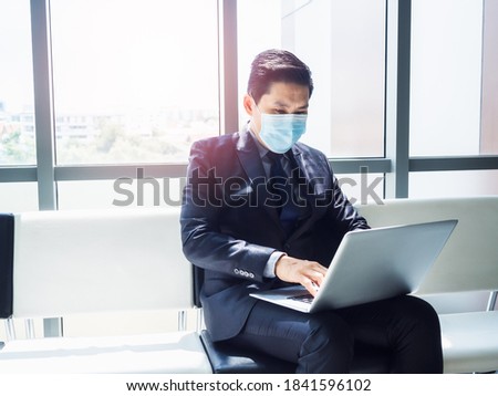 Asian businessman in suit wearing protective face mask using laptop computer on his lap while sitting in modern office building near huge glass window.