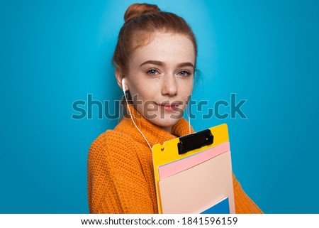 Close up photo of a ginger student with freckles posing with folders and earphones on a blue studio wall in a yellow sweater