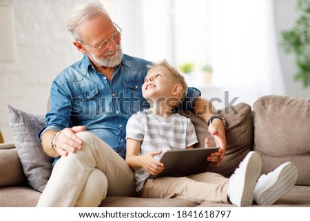 Happy family senior grandfather and boy sitting on sofa and watching cartoon on tablet on weekend day at home together

