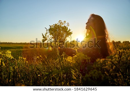 Beautiful woman with long hair in red dress holding bouquet of yellow flowers and enjoying summer sunny day in green field with green grass in sunset with nice orange sun