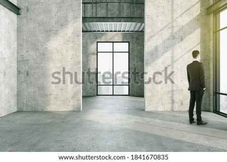 Businessman standing in modern concrete interior and copy space on gray wall. Performance and presentation concept. Mock up