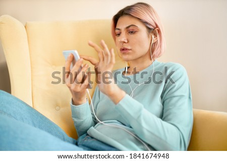 Portrait of beautiful stylish young female with pinkish hair sitting comfortably in armchair with cell phone, typing text message or uploading new pic via social network, wearing earphones