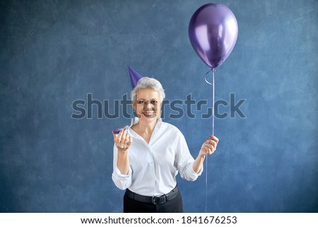 Waist up picture of attractive successful European mature businesswoman in formal elegant clothes and conical hat eating dessert at corporate party, holding helium balloon, smiling happily