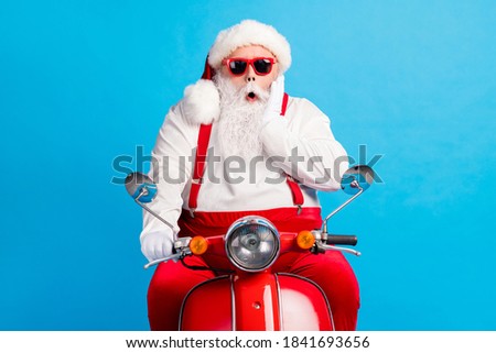 Photo of grandfather grey beard ride retro scooter palm cheekbone open mouth wear santa claus x-mas costume suspenders sunglass white shirt cap gloves isolated blue color background
