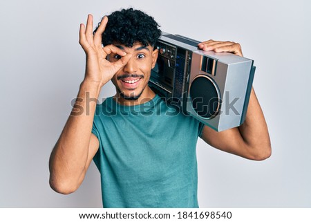 Young arab handsome man holding boombox, listening to music smiling happy doing ok sign with hand on eye looking through fingers 