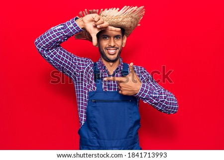 Young latin man wearing farmer hat and apron smiling making frame with hands and fingers with happy face. creativity and photography concept. 