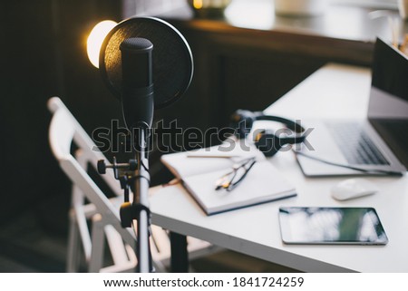 Items for recording podcast: professional microphone, earphones and laptop on white table in cozy home studio with black walls and lots of plants.