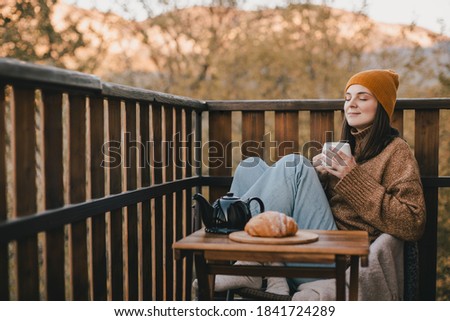 Young woman in knitted sweater and hat drinking tea and eating fresh croissants on cozy balcony of a wooden country house on autumn day.  Royalty-Free Stock Photo #1841724289