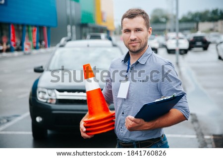 Male driving instructor standing with orange traffic cones and clipboard in his hands. Caucasian man working in driving school. Blurred gray car in background. Royalty-Free Stock Photo #1841760826