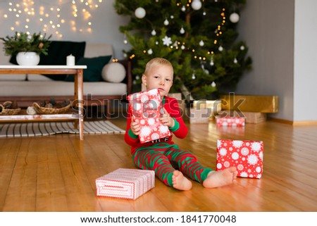 Little boy and a girl in cozy pajamas and Santa Claus hats unpack gifts at home in the new year's interior.