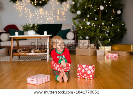 Little boy and a girl in cozy pajamas and Santa Claus hats unpack gifts at home in the new year's interior.