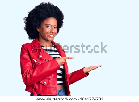 Young african american girl wearing red leather jacket amazed and smiling to the camera while presenting with hand and pointing with finger. 