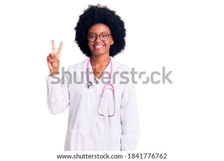 Young african american girl wearing doctor coat and stethoscope smiling looking to the camera showing fingers doing victory sign. number two. 