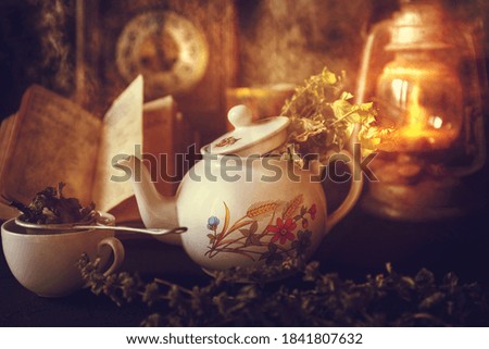 Cup of Tea and Teapot on wooden table with vintage clock, burning lantern and old book in the background