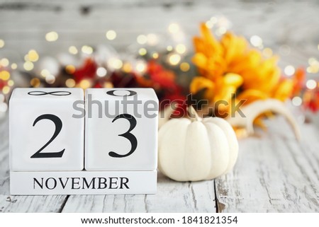 White wood calendar blocks with the date November 23rd and autumn decorations over a wooden table. Selective focus with blurred background. 