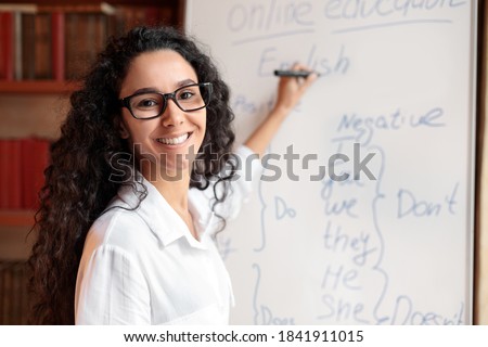 Education And Learning Concept. Portrait of smiling female teacher standing at whiteboard, explaining grammar rules to students. Excited woman in glasses looking at camera, writing on the board Royalty-Free Stock Photo #1841911015