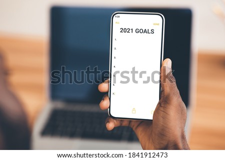 2021 Goals List. Black Businessman Holding Smartphone With Goal Checklist For Upcoming New Year Planning Business Projects And Life Sitting At Laptop Indoors. Closeup, Selective Focus, Cropped Royalty-Free Stock Photo #1841912473
