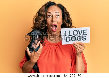 Middle age hispanic woman holding chihuahua dog and paper with i love dogs phrase afraid and shocked with surprise and amazed expression, fear and excited face. 