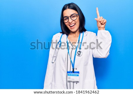 Young beautiful latin woman wearing doctor stethoscope and id card pointing finger up with successful idea. exited and happy. number one. 