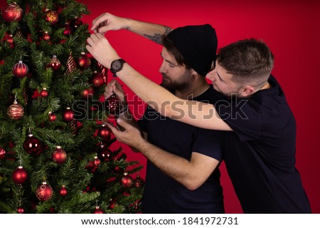 Young male homosexual couple decorating xmas tree together celebrating New Year, two stylish gay men preparing for family party. Merry Christmas, winter holiday concept. Red background