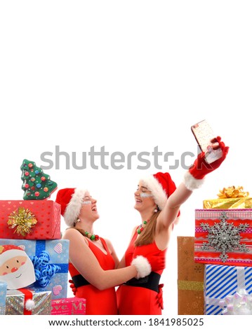 Two happy teenage girls dress up as Santa Claus at 
Christmas time and stand between Christmas presents. 
They cheerfully take selfies of each other with their 
cell phone. 