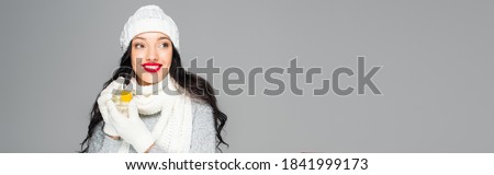 happy woman in winter outfit holding bottle with perfume isolated on grey, banner