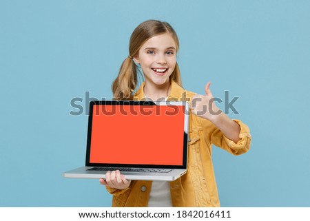 Funny little kid girl 12-13 years old in yellow jacket isolated on blue background. Childhood lifestyle concept. Mock up copy space. Hold laptop pc computer with blank empty screen, showing thumb up