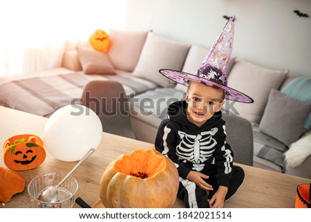 Halloween, holiday and childhood concept - Smiling little boy in party costume of skeleton with jack-o-lantern pumpkin sitting on table and having fun at home