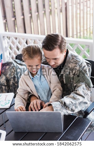 Military serviceman hugging girl and looking at laptop, while sitting at table