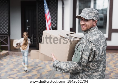 Happy military serviceman holding cardboard box and looking at camera with blurred girl on background