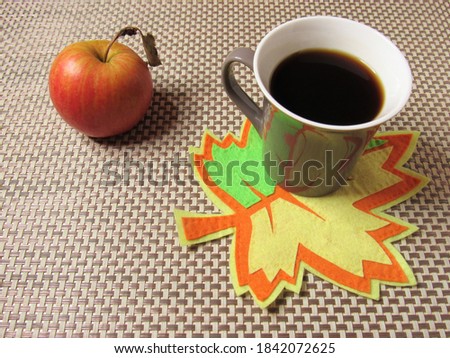 On the table is a cup of coffee and an apple. The cup stands on a bright maple leaf. This autumn maple leaf is made of felt.