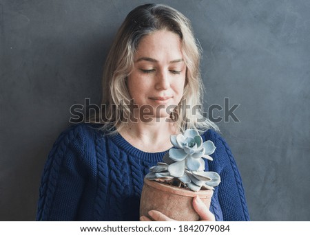 Woman holding a succulent plant on a pot, looking down at it, wearing a blue knitted sweater on a dark gray wall background