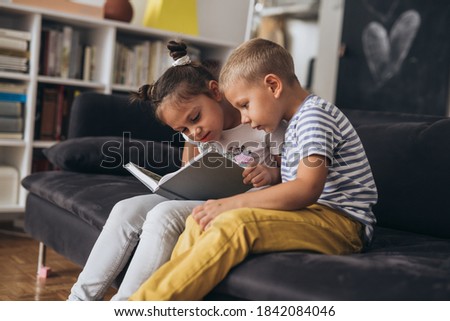 brother and sister sitting on sofa and reading book at home