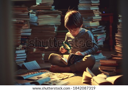 Cute little boy with a magnifier is examining herbarium among the piles of books. Image with selective focus and toning Royalty-Free Stock Photo #1842087319