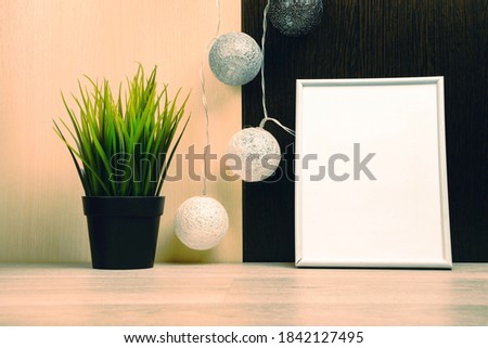 White frame with place for text and a green plant in a black pot against the background of a black and white wall and decorative balls. Stylish room interior.