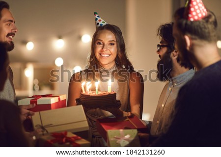 Thank you for the surprise, guys. Portrait of happy young woman in her twenties surrounded by friends, holding her birthday cake with burning candles and looking at camera during party at home or cafe