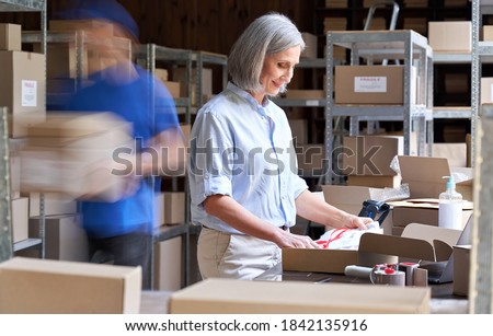 Older female small business owner worker packing post shipping ecommerce retail order in box in warehouse with courier hurry in blur motion to deliver parcels. Fast speed express rush delivery concept Royalty-Free Stock Photo #1842135916