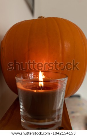 Close-up photo of an organic pumpkin in focus with an aromatic candle located on top of a wooden shelf. Pumpkins are the main symbol of autumn and Halloween.