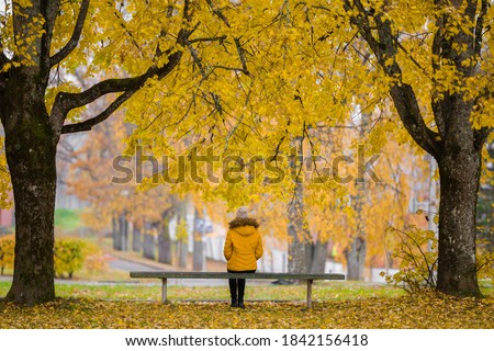 Young woman in yellow jacket sitting alone on bench between trees at beautiful day at park. Golden autumn. Thinking about life. Spending time alone in nature. Peaceful atmosphere. Back view. Royalty-Free Stock Photo #1842156418