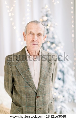 Close up of senior man in checkered jacket and light shirt, posing to camera at cozy room at home over christmas tree background. Christmas, winter holidays, elderly age and people concept