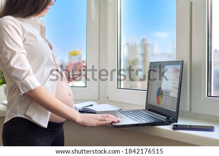 Young pregnant business woman working at home with laptop, drinking mineral water with lemon in glass, window background. Remote work at home, pregnancy and business