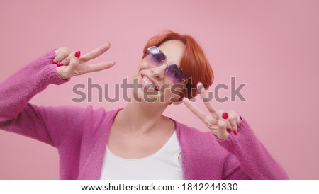 Beautiful woman in 40s with pink heart shaped glasses showing peace sign with both hands. Isolated on pink purple background. 