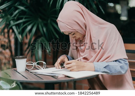 Muslim female student taking down note in diary. Concentrated girl studying on a book and taking down note sitting at the desk.