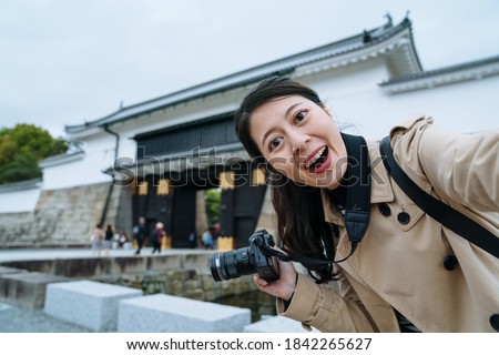 happy young lady holding camera is taking a selfie with smartphone at the entrance of Nijo Castle. asian female traveler facing to camera is excited to see the famous historic building in Japan.