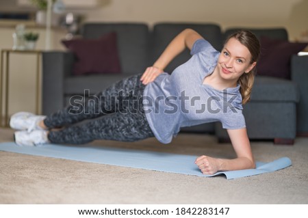 Young fit woman in sportswear looks at camera and doing yoga exercise on a mat at home. Concept of training in living room. Stay at home activities. Healthy lifestyle.