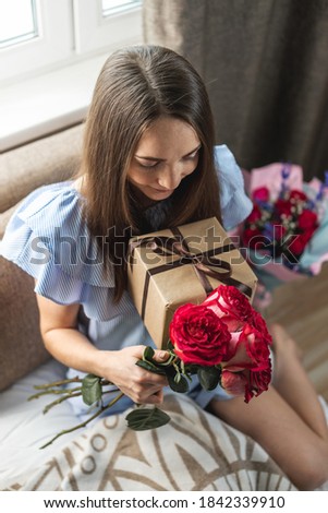 Happy young woman with a beautiful bouquet of fresh roses and a gift box in her hands at home. Concept of a holiday, romance, good and cozy mood in birthday.