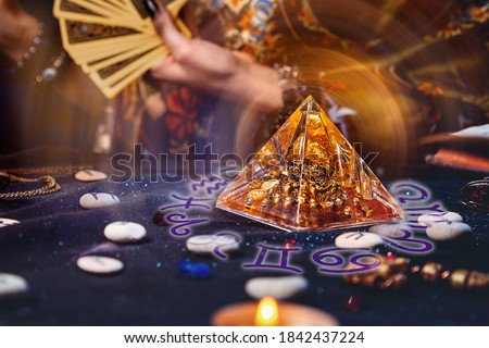 Magic divination and esotericism. Magic glass pyramid with a magical glow. In the background, a fortune teller holds a fan of Tarot cards. Close-up of hands Royalty-Free Stock Photo #1842437224