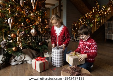 Excited small brother and sister kids sit on floor in living room near Christmas tree unpack presents on Christmas Eve. Happy little kids have fun unwrap open gifts on New Year in cozy home.