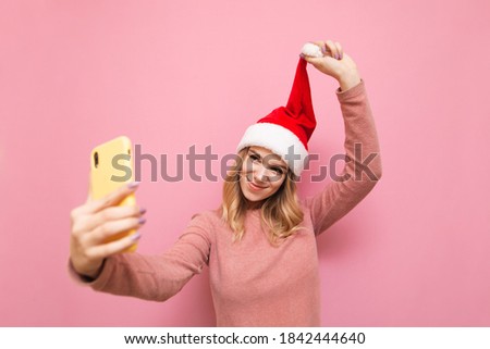 Portrait of happy woman in christmas hat, isolated on pink background, taking selfie on smartphone camera and smiling. Smiling lady posing on smartphone camera with happy face, showing santa hat.